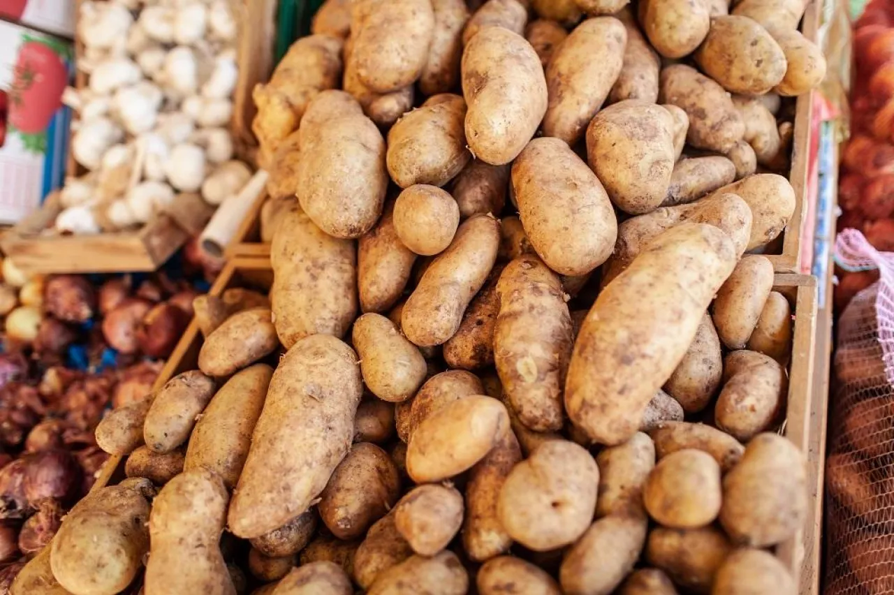 A closeup of potatoes of different shapes and sizes in wooden boxes. There are other vegetables like onions and garlic on the blurred background