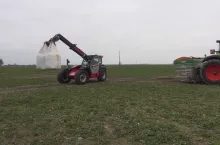 Ładowarka teleskopowa z uniesionym workiem nawozu i ciągnik z rozsiewaczem na dużym polu rolnym. Maszyny rolnicze pracują w terenie, przygotowując uprawy do nowego sezonu wegetacyjnego. W tle widać płaską, otwartą przestrzeń z pojedynczymi drzewami i pochmurnym niebem