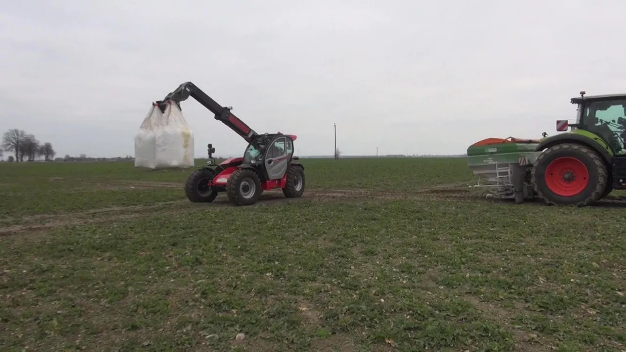 Ładowarka teleskopowa z uniesionym workiem nawozu i ciągnik z rozsiewaczem na dużym polu rolnym. Maszyny rolnicze pracują w terenie, przygotowując uprawy do nowego sezonu wegetacyjnego. W tle widać płaską, otwartą przestrzeń z pojedynczymi drzewami i pochmurnym niebem